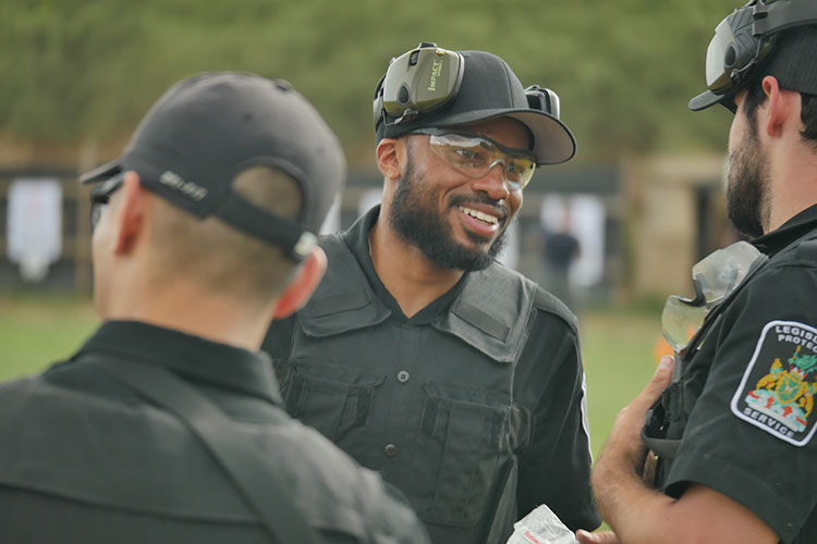 officer standing in between two officers, smiling with noise cancellation earmuffs propped on top head